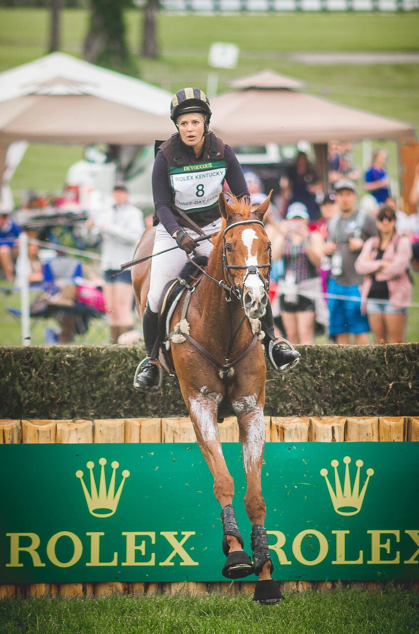 Rider on horse, completing a jump over a hedge fence with Rolex logos underneath and people looking on.