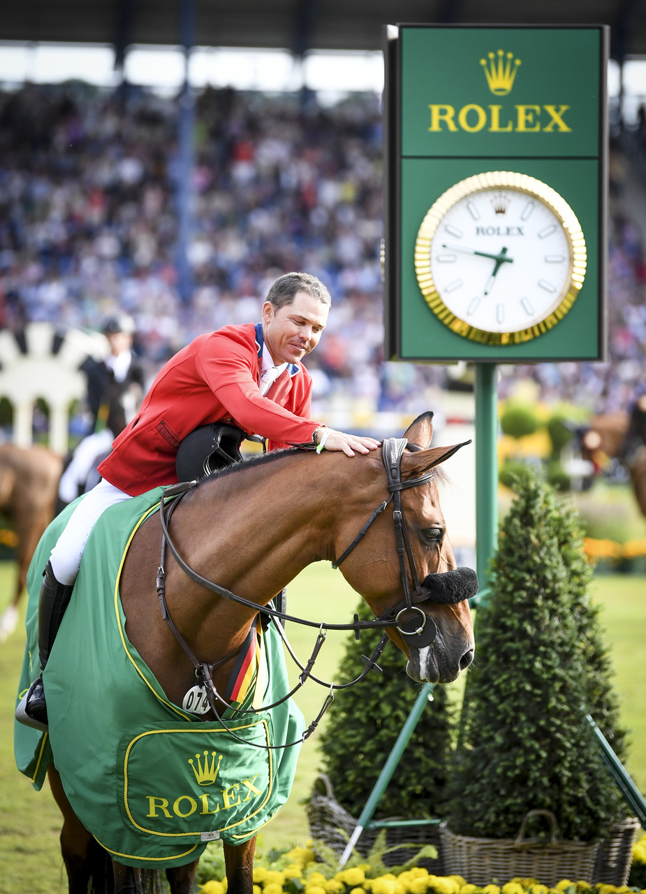 Rider in a red jacket, holding helmet, patting the horses neck. Rolex clock in background, Rolex jacket around the horse. In a stadium of people.
